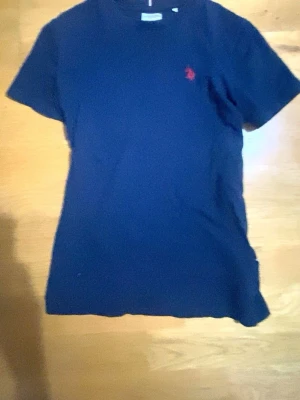 Marinblå t-shirt från U.S. Polo Assn. - Snygg marinblå t-shirt från U.S. Polo Assn. med klassisk röd logga broderad på bröstet. T-shirten har rund halsringning och korta ärmar, perfekt för en clean och stilren look. Tillverkad i mjuk bomull för skön känsla hela dagen.
