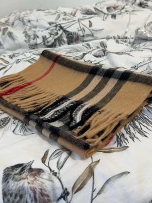 Burberry halsduk - Säljer en burberry halsduk med rutigt mönster i svart, vitt och rött. Halsduken har fransar i kanterna och är mjuk och skön, perfekt för kyliga dagar. Passar till de flesta outfits och ger en tidlös look.
