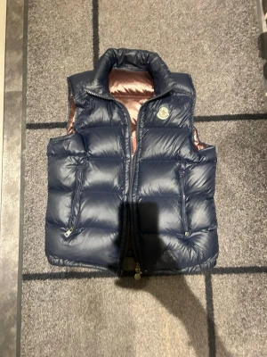 ÄKTA Mörkblå dunväst från Moncler - Snygg mörkblå dunväst äkta köpt från Moncler med hög krage och två dragkedjeförsedda fickor framtill. Västen har quiltad design och klassisk Moncler-logga på bröstet. Insidan är glansigt brun. Perfekt för lager-på-lager under kyliga dagar.