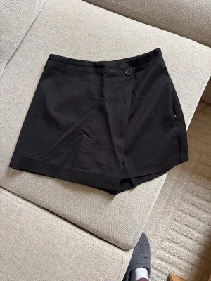 Svarta skorts från H&M Divided - Säljer ett par svarta skorts från H&M Divided i storlek 34. De har omlott-design framtill med knapp i midjan och är gjorda i ett mjukt bomullsliknande material. Perfekta för dig som vill ha shortskomfort men kjol-look.