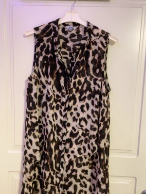 Ärmlös leopardmönstrad klänning - Säljer en ärmlös klänning med leopardmönster i svart och beige. Klänningen har knappar framtill och en avslappnad passform. Perfekt för dig som vill sticka ut med ett djurmönster. Märke: Gina Tricot.