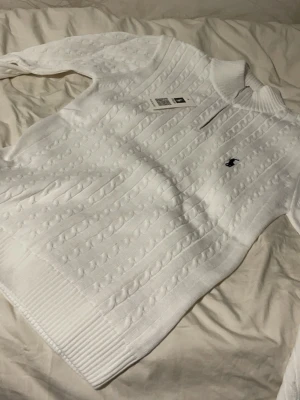 Vit kabelstickad tröja Polo Ralph Lauren - Kabelstickad vit tröja från Polo Ralph Lauren med klassisk rund halsringning och ribbade muddar. Tröjan har den ikoniska lilla broderade loggan på bröstet och är tillverkad i mjukt material. Perfekt för en clean och stilren look.