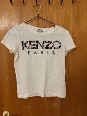 Vit Kenzo t-shirt med logga - Snygg vit t-shirt från Kenzo med stort tryck på bröstet där det står 'KENZO PARIS' i svart och rosa. Klassisk passform och rund halsringning. Perfekt för dig som gillar streetwear och vill ha något trendigt i garderoben.
