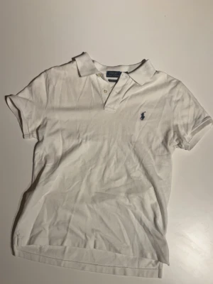 Vit pikétröja från Polo Ralph Lauren - Klassisk vit pikétröja från Polo Ralph Lauren i bomull med blå broderad logga på bröstet. Modellen är Classic Fit med korta ärmar, krage och två knappar framtill. Perfekt basic till garderoben och enkel att styla.