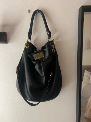 Svart handväska Marc by Marc Jacobs - Snygg svart handväska från Marc by Marc Jacobs med guldfärgade metalldetaljer och präglad logoplatta framtill. Väskan är tillverkad i mjukt skinn och har både handtag och axelrem för flexibel användning. Perfekt för dig som vill ha en klassisk och stilren accessoar.