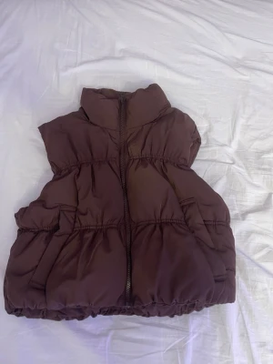 Brun dunväst från H&M Divided - Snygg brun dunväst från H&M Divided med hög krage och dragkedja framtill. Västen har två fickor på sidorna och en puffig, croppad passform som ger en trendig look. Perfekt att slänga över en hoodie eller tröja för extra värme.