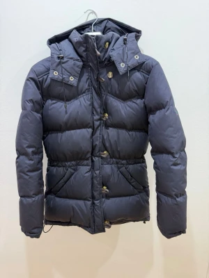 Marinblå dunjacka med huva  - Snygg marinblå dunjacka med huva och dragkedja samt knappar framtill. Jackan har quiltad design, långa ärmar och praktiska fickor. Perfekt för kalla dagar och har en modern, puffig look.