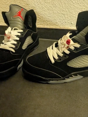 Nike Air Jordan 5 Retro svart/grå - Snygga Nike Air Jordan 5 Retro sneakers i svart mocka med vita snören och grå detaljer. Skorna har den klassiska Jumpman-loggan på plösen och genomskinlig sula med Nike- och Jumpman-märke undertill. Perfekta för dig som gillar streetwear och ikoniska sneakers.