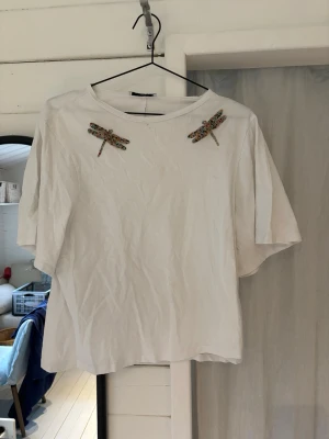 Vit t-shirt med pärlbroderade trollsländor Mango - Vit t-shirt från Mango med lite längre armar och rund hals. På axlarna sitter två färgglada pärlbroderade trollsländor som ger en unik touch. 