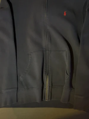 Mörkblå hoodie från Polo Ralph Lauren - Mörkblå hoodie med dragkedja från Polo Ralph Lauren. Klassisk design med röd broderad logga på bröstet, känguruficka framtill och mjukt fleecefoder. Perfekt för en chill och stilren look. Materialet är bomullsmix som känns skönt mot huden.