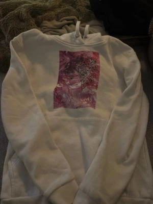 Vit hoodie med rosa anime-tryck - Säljer en vit hoodie med ett stort rosa anime-inspirerat tryck på bröstet. Hoodien har huva med dragsko och en klassisk känguruficka framtill. Perfekt för dig som gillar streetwear och unika prints.