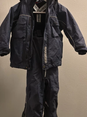 Polarn O. Pyret mörkblå täckbyxor med hängslen och vinterjacka till barn strl 92 - Säljer ett par mörkblå täckbyxor och matchande jacka med avtagbar fleecetröja från P.O.P  med justerbara hängslen och reflexdetaljer. Byxorna har dragkedja framtill, perfekta för kalla dagar utomhus. 