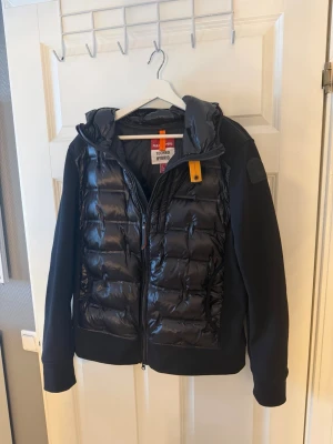 Svart pufferjacka från Parajumpers - Snygg svart pufferjacka från Parajumpers med quiltad front och huva. Jackan har dragkedja, ribbade muddar och en gul detalj vid halsen. Materialmix med glansig polyester framtill och mjuka ärmar. Perfekt för kalla dagar.
