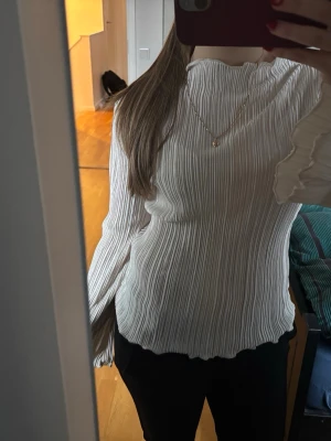 Vit plisserad blus med volangärm - Snygg vit plisserad blus med hög hals och långa volangärmar. Blusen har en luftig passform och vågig kant nertill och vid ärmslut. Perfekt att styla med både jeans och kjol för en trendig look.
