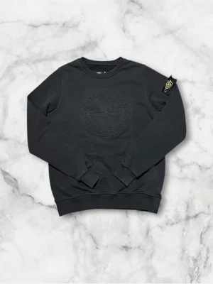 Mörkblå sweatshirt från Stone Island - Mörkblå sweatshirt från Stone Island med rund halsringning och ribbade muddar. Stort broderat Stone Island-emblem på bröstet och klassisk logotyp-patch på vänster ärm. Perfekt för en clean och stilren streetwear-look.