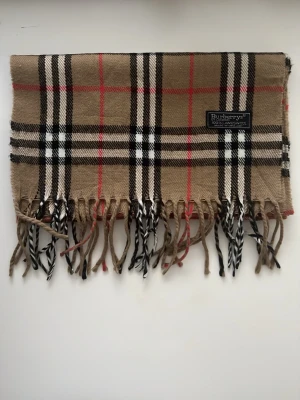 Rutig ullväst från Burberrys - Klassisk väst från Burberrys i 100% lammull med ikoniskt rutigt mönster i beige, svart, vitt och röda detaljer. Västen har fransar nedtill och en mjuk, varm känsla. Perfekt för dig som vill ha en tidlös och snygg look.