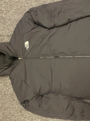 Svart pufferjacka från The North Face, barnstorlek kanske 158-164 - Säljer en svart pufferjacka från The North Face med huva och dragkedja framtill. Jackan har en clean look med diskret logga på bröstet och är perfekt för kalla dagar. Tillverkad i slitstark polyester och har en klassisk, lite oversized passform.