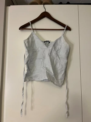 Ljusblå omlottopp med knytband - Säljer en ljusblå linnetopp med smala axelband. Toppen har långa knytband i sidorna som kan knytas i midjan för en snygg passform. Perfekt till sommaren och enkel att styla med jeans eller kjol.