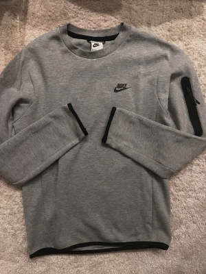 Grå långärmad Nike tech sweatshirt - Säljer en stilren grå Nike tech sweatshirt (ÄKTA) med svart logga på bröstet och en praktisk ficka med dragkedja på ena ärmen. Tröjan har svarta detaljer vid ärmslut och nederkant. Perfekt för chill eller träning.