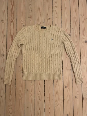 Beige kabelstickad tröja Polo Ralph Lauren - Kabelstickad tröja i beige från Polo Ralph Lauren med rund halsringning och den klassiska mörkblå logotypen broderad på bröstet. Tröjan har långa ärmar och ribbade muddar vid ärmslut och nederkant. Perfekt för en stilren och avslappnad look. Mycket bra skick. Nästan oanvänd. Bara att skriva vid frågor