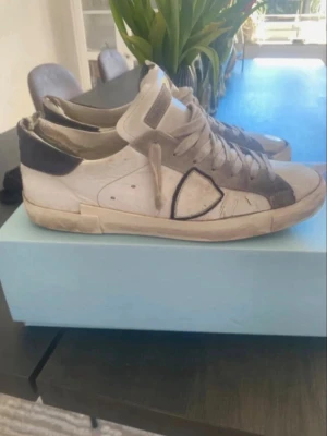 Philippe Model vita sneakers - Säljer ett par vita Philippe Model sneakers med svarta detaljer och beige inslag. Skorna har snörning, rund tå och platt sula. Tillverkade i skinn med gummisula och har den klassiska logotypen på sidan. Perfekta för en avslappnad och trendig stil.