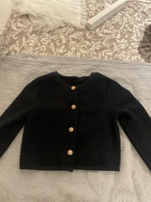 Svart cardigan jacka från Gina Tricot - Stilren svart cardigan jacka från Gina Tricot med rund halsringning och dekorativa guldknappar framtill. Jackan är i ett mjukt, stickat material med en lätt struktur och har lång ärm. Perfekt för att lyfta vilken outfit som helst.