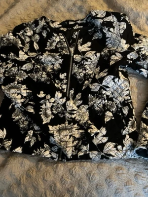 Svartvit blommig bomberjacka Divided - Snygg svart bomberjacka från HM med vitt blommönster och dragkedja framtill. Jackan har klassisk bomberform och är perfekt för dig som vill sticka ut med en cool och unik look. Passar till många olika outfits och har lång ärm.