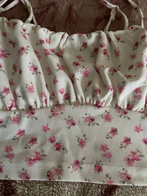 Blommig vit linnetopp med smala band - Supersöt linnetopp i vitt med små rosa blommor och smala axelband. Toppen har en lätt rynkad detalj framtill och är croppad i modellen. Perfekt för varma dagar och ger en romantisk vibe till din outfit.