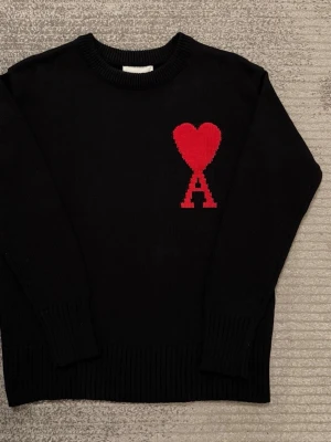 Svart stickad tröja  - Svart stickad tröja med ikoniskt rött hjärta och bokstaven A broderat på bröstet. Tröjan har rund halsringning, ribbade muddar och nederkant. Perfekt för dig som gillar stilrena plagg med en twist. Helt oanvänd.