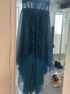 Blå glittrig långklänning - Säljer en blå  långklänning med glittrande detaljer och transparent tyll över kjolen. Klänningen har en asymmetrisk nederkant och markerad midja. Perfekt för dig som vill sticka ut på bal eller fest.