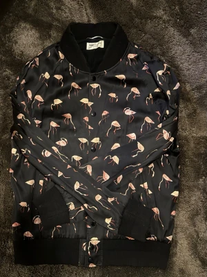 Svart bomberjacka från Saint Laurent -  Äkta Saint Laurent Paris bomberjacka. Flamingomönster, svart/rosa, tryckknappar. Storlek 48 (M), unisex. Mycket gott skick. Pris satt för snabb försäljning.