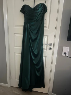Mörkgrön långklänning Dressroom Linné - Elegant mörkgrön långklänning från Dressroom Linné i glansigt tyg. Klänningen har offshoulder-modell, draperad överdel och markerad midja med ett snyggt fall. Perfekt för bal eller festliga tillfällen. Levereras i original klänningspåse med prislapp kvar. Originalpris: 4500kr