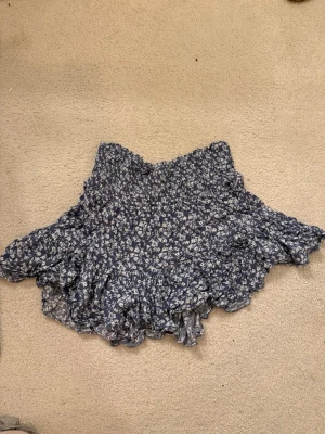 Blommig volangkjol från 157 - Supersöt kort kjol från 157 med blå botten och vitt blommönster. Kjolen har bred smock i midjan och flera lager volanger som ger en flowy känsla. Perfekt för dig som gillar en feminin och avslappnad stil.