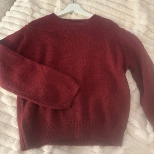 Mörkröd stickad tröja i ullmix - Mysig mörkröd stickad tröja med rund halsringning och långa ärmar. Tröjan har ribbstickade muddar vid ärmslut och nederkant, vilket ger en klassisk och avslappnad look. Perfekt för kyliga dagar och enkel att matcha med jeans eller kjol.