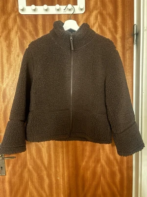 Brun teddyjacka från Gina Tricot - Mysig brun teddyjacka från Gina Tricot med hög krage och hel dragkedja framtill. Jackan har en loose passform och är tillverkad i mjukt teddy-material som håller dig varm under kyliga dagar. Perfekt för en avslappnad och trendig look. Den är i ett bra skick, bara att de finns lite sminkfläckar på kragen. Dom ser ni på bilden där uppe, annars är jacka super fin och bra skick