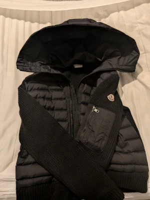 Moncler cardigan  - Snygg svart dunväst från Moncler med stickade detaljer och stor huva. Västen har en dragkedja framtill, en bröstficka med dragkedja och Moncler-log sopatch på bröstet. Perfekt för lager-på-lager och streetstyle.