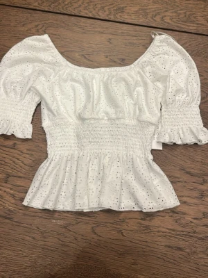 Vit blus med broderade detaljer - Supersöt vit blus från Kirious med broderade hål och volangkant. Modellen har puffiga korta ärmar med smock och bred smockad midja som ger en feminin siluett. Helt ny med lapp kvar💕