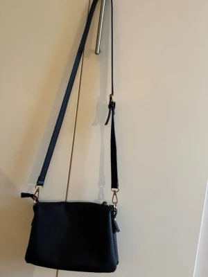 Svart axelväska i skinnimitation - Snygg svart axelväska med justerbart axelband och guldfärgade detaljer. Väskan har en enkel, stilren design med dragkedja upptill och en extra ficka med dragkedja på baksidan. Har använts några gånger och dragkedjan är en aning seg men den funkar som den ska skulle jag säga .