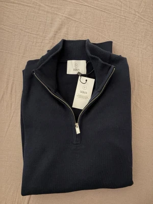 Mörkblå halvzip-tröja från Bläck - Snygg mörkblå stickad tröja från Bläck med halvzip och hög krage. Tröjan har ribbade muddar och är tillverkad i mjukt material som passar perfekt för lager på lager. Enkel och stilren design som funkar till det mesta.