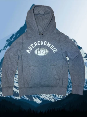 Abercrombie & fitch M - Snygg grå hoodie från Abercrombie med broderad logga och siffran 92 framtill. Klassisk känguruficka och dragsko i huvan. Tillverkad i mjukt bomullsmaterial som är skönt mot huden. Perfekt för en avslappnad och sportig stil.
