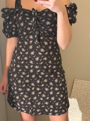 Svart blommig kortklänning med puffärm - Supersöt svart kortklänning med små blommor i vitt och rosa. Klänningen har puffärmar, snörning framtill och en smickrande v-ringning. Perfekt för sommarens alla tillfällen och ger en romantisk vibe.