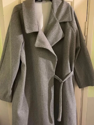 Grå kappa - Stilren grå kappa med bred krage och knytbälte i midjan. Långärmad och rak modell som ger en clean och modern look. Perfekt för dig som gillar minimalistisk stil och vill ha en jacka som funkar till många outfits.