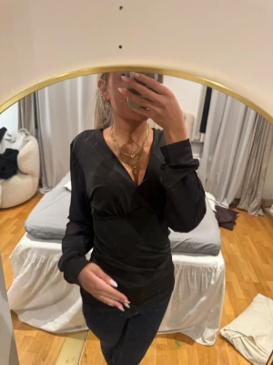 Svart omlottopp med v-ringning - Svart omlottopp med djup v-ringning och långa ärmar i ett lätt, transparent material. Sitter figurnära och har en stilren look som passar till många outfits. Perfekt för dig som gillar trendiga och enkla plagg.