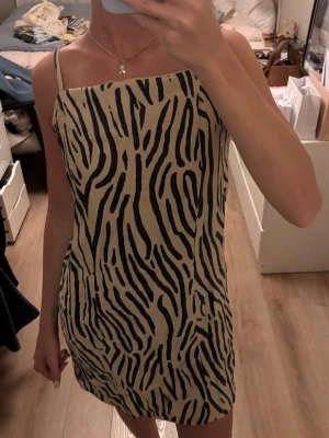 Beige zebramönstrad kortklänning - Trendig kort klänning med beige bas och svart zebramönster. Klänningen har smala axelband och rak halsringning, perfekt för dig som vill sticka ut. Snygg och enkel att styla med accessoarer.