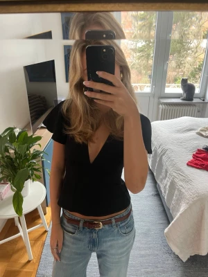 Svart v-ringad blus med knyt i ryggen - Snygg svart topp med djup v-ringning och korta ärmar. Toppen har band i sidan som man knyter som en rosett i ryggen. Köptes på Gina Tricot för ca ett år sen och är i nyskick. 