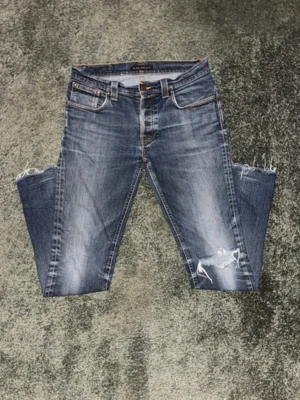 Feta Blå grim Tim’s från Nudie Jeans - Säljer ett par blå jeans från Nudie Jeans med feta slitningar och hål på knäna. Klassisk femficksmodell med orange sömmar och Nudie-logga på bakfickan. Jeansen har en rak passform och är tillverkade i bomull. Det står size 34/34 men de passar ärligt 31/32 vet inte varför dock. Midjan är 42 cm och längden är runt 104 cm. Priset kan diskuteras, skickar snabbt✌️💯