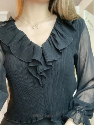 Svart volangblus från Zara - Trendig svart blus från Zara med volangdetaljer längs V-ringningen och framtill. Blusen är gjord i ett tunt, transparent tyg med plisserad struktur och har långa ärmar med resår och volang vid ärmslut. Perfekt för dig som gillar en chic och feminin stil. Använd väldigt få gånger. 🩷