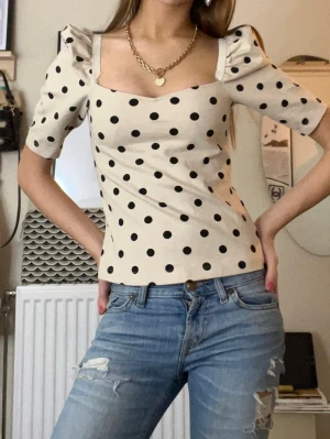 Prickig puffärm topp i beige och svart - Säljer en beige topp med svarta prickar och puffärmar. Toppen har en rak passform, fyrkantig urringning och halvlånga ärmar. Går att bära både offshoulder och med ärmar, toppen har en trendig vintage-vibe. Perfekt att styla med jeans eller kjol.