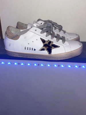 Vita golden goose sneakers med leopardstjärna original pris runt 9000 - Snygga vita sneakers från Golden Goose med grå snörning, beige detaljer och en stjärna i leopardmönster på sidan. Skorna har låg profil och platt sula, använda en gång, säljer pga att jag köpte fel storlek