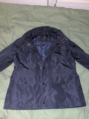 Mörkblå vindjacka från Bondelid - Snygg mörkblå vindjacka från Bondelid med huva och flera praktiska fickor framtill. Jackan har tryckknappar och dragkedja, samt en lätt glansig finish. Perfekt för blåsiga dagar och enkel att matcha med olika outfits. Kvitto finns med har använt jackan 1 gång pris går att dissekutera vid snabb affär 